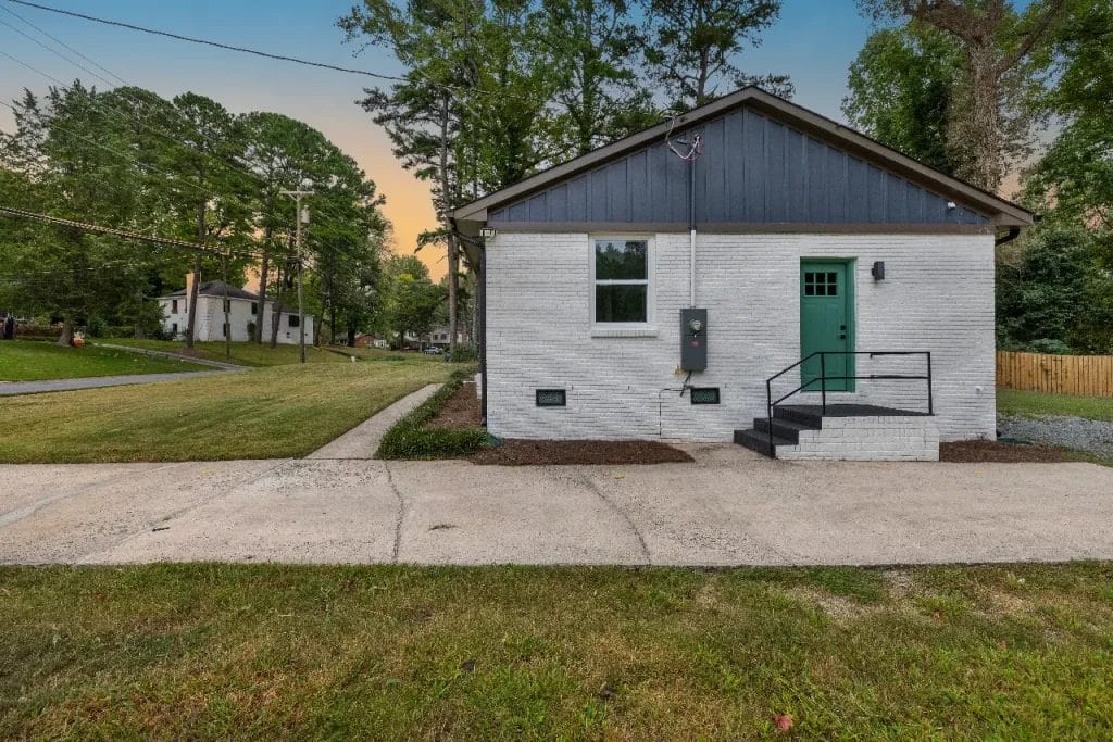 Small white brick house with a green front door, black trim, and steps leading to the entrance. The house sits on a grassy lot with trees and a paved sidewalk in front, at sunset.