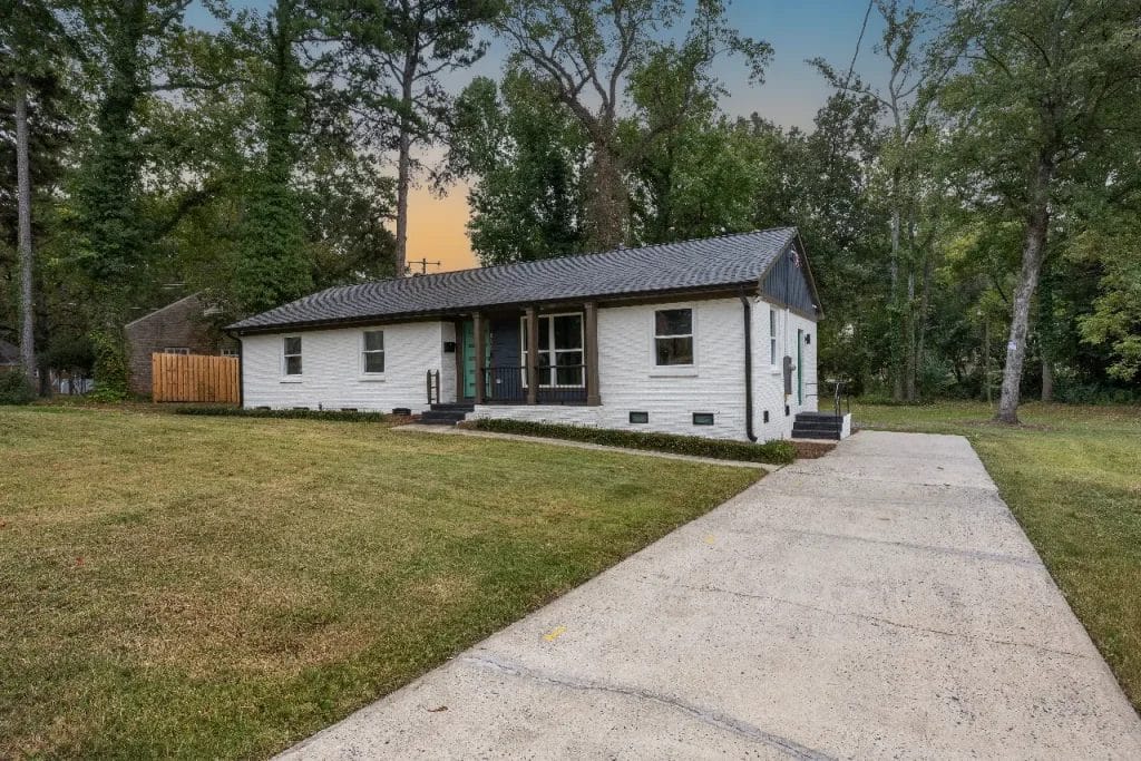 Single-story white brick house with a dark roof, black trim, and front porch, set on a large grassy yard with a concrete driveway, surrounded by tall trees at dusk.