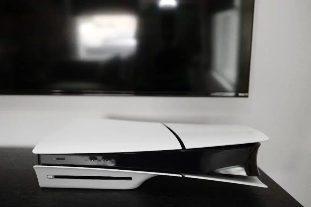 A PlayStation 5 gaming console sits horizontally on a black surface in front of a large, wall-mounted flat-screen TV. The photo is focused on the PS5, with the TV slightly blurred in the background.