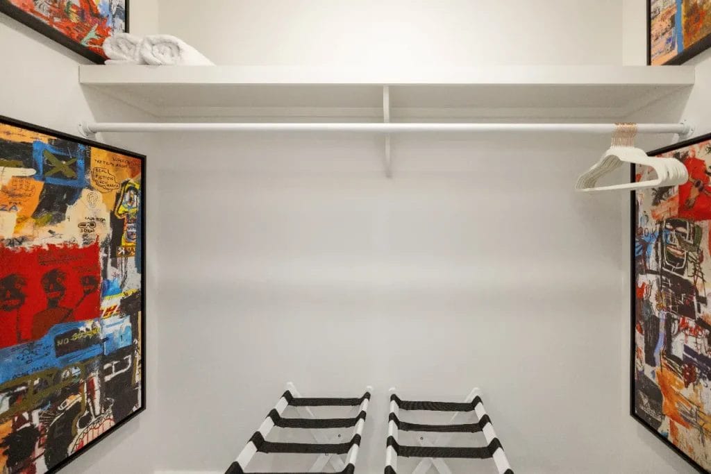 An empty walk-in closet with a single shelf holding a folded towel, a hanging rod with a few white hangers, two luggage racks, and colorful abstract art on the side walls. The space is brightly lit.
