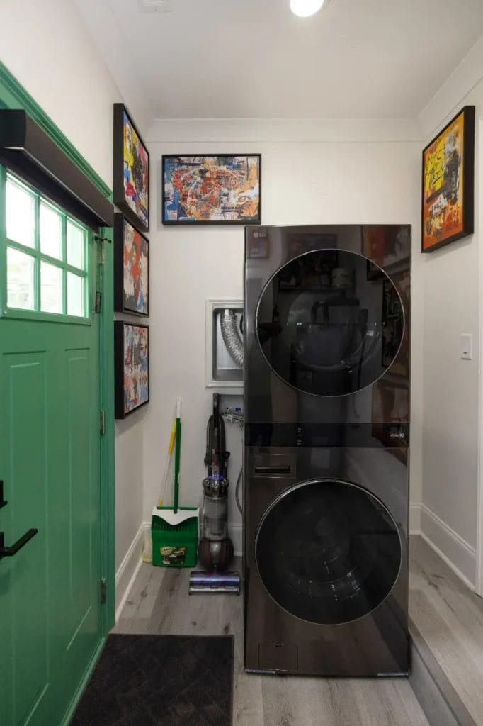 A compact laundry area with a stacked washer and dryer beside a green door, colorful artwork on the walls, and cleaning supplies including a vacuum, broom, and mop in the corner.