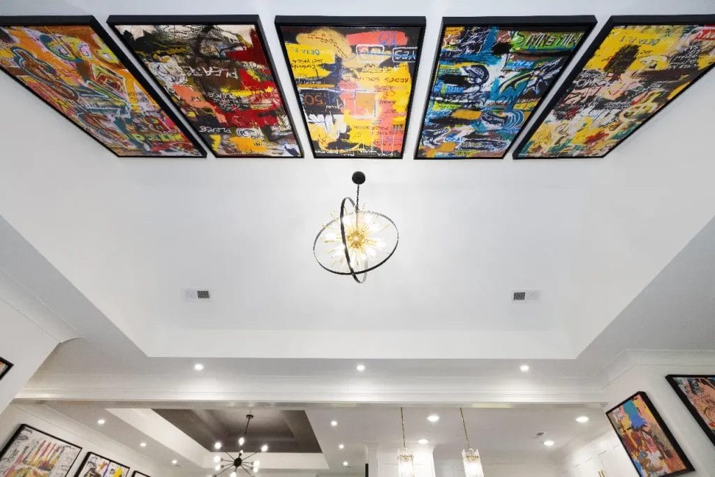 Colorful abstract paintings are displayed on the ceiling of a modern white room with recessed lighting and a central decorative chandelier. More art and lighting fixtures are visible on the walls and ceiling below.