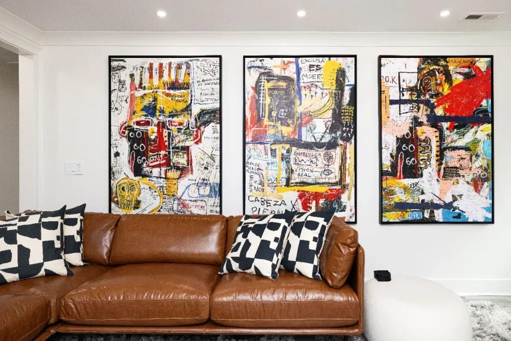 A modern living room with a brown leather sofa, black-and-white patterned pillows, and three large abstract art pieces with bold colors and text hanging on a white wall above the couch.