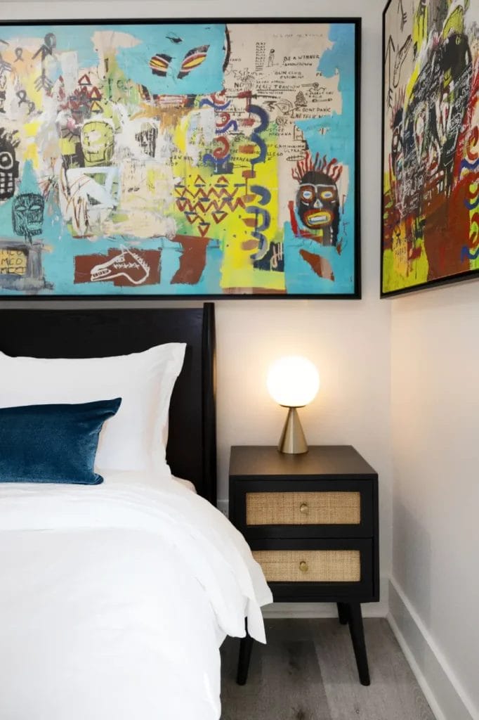 A modern bedroom with a white bed, dark headboard, and blue pillow. A black nightstand with two drawers and a round lamp sits beside the bed. Colorful abstract art hangs above the bed and nightstand.