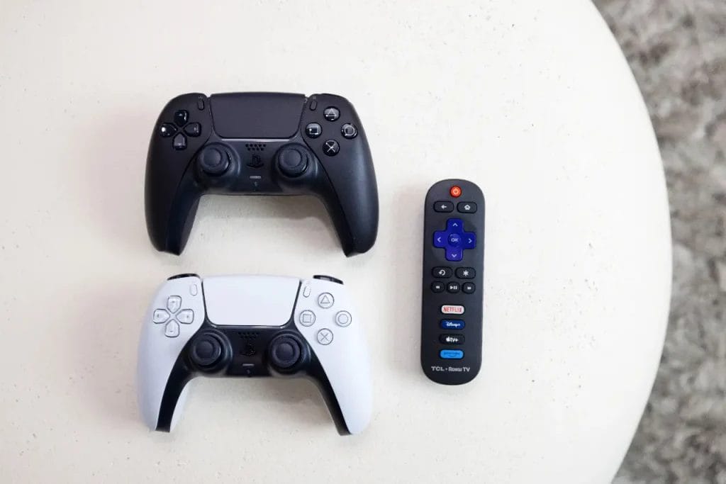 Two video game controllers, one black and one white, are placed side by side above a small black TV remote with blue directional buttons on a light-colored surface.