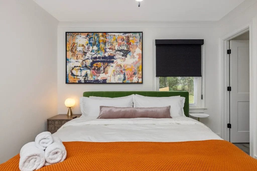 A neatly made bed with white linens, an orange blanket, and a green headboard, topped with a folded towel. Above the bed hangs an abstract painting, and a window with a black shade is on the right.