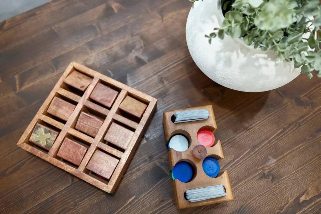 A wooden tic-tac-toe game and a wooden sorting toy with colored discs are placed on a wooden table next to a white vase with green foliage.