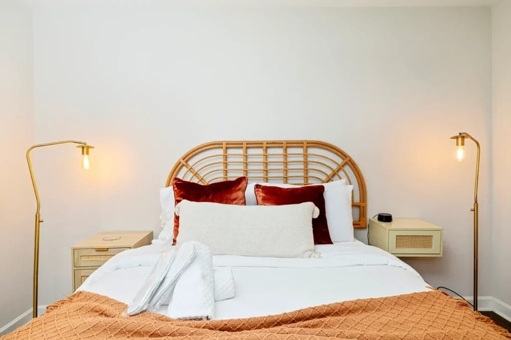 A neatly made bed with a rattan headboard, rust-colored and white pillows, and a rust-colored blanket. Two bedside tables with lamps and a folded white towel are on the bed, all against a white wall.