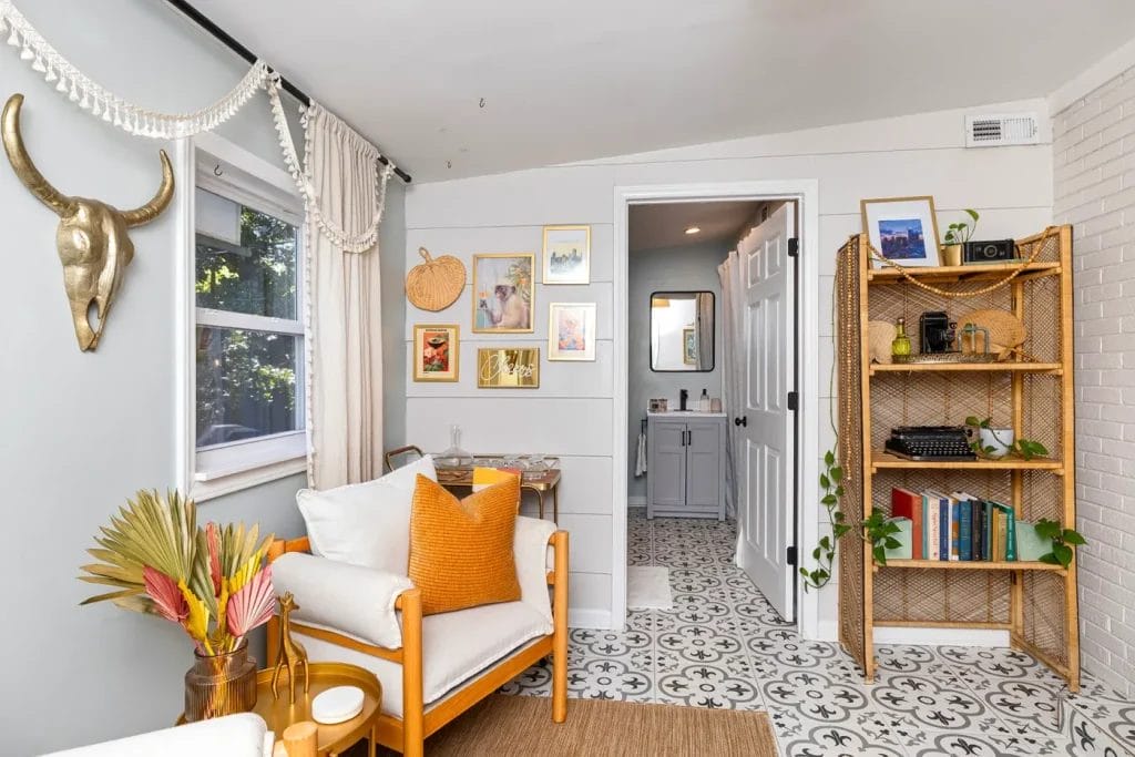 A cozy living space with a white armchair, orange pillow, and decorative plants. Art and a gold bull skull hang on the walls. In the background, a small table, shelving with books, and a view into a patterned-tile bathroom are visible.