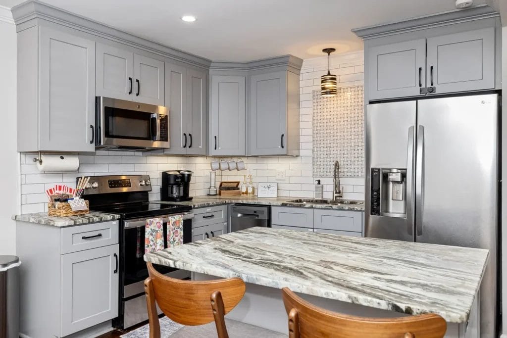 Modern kitchen with gray cabinets, stainless steel appliances, marble countertops, a kitchen island with two wooden chairs, white subway tile backsplash, and under-cabinet lighting.