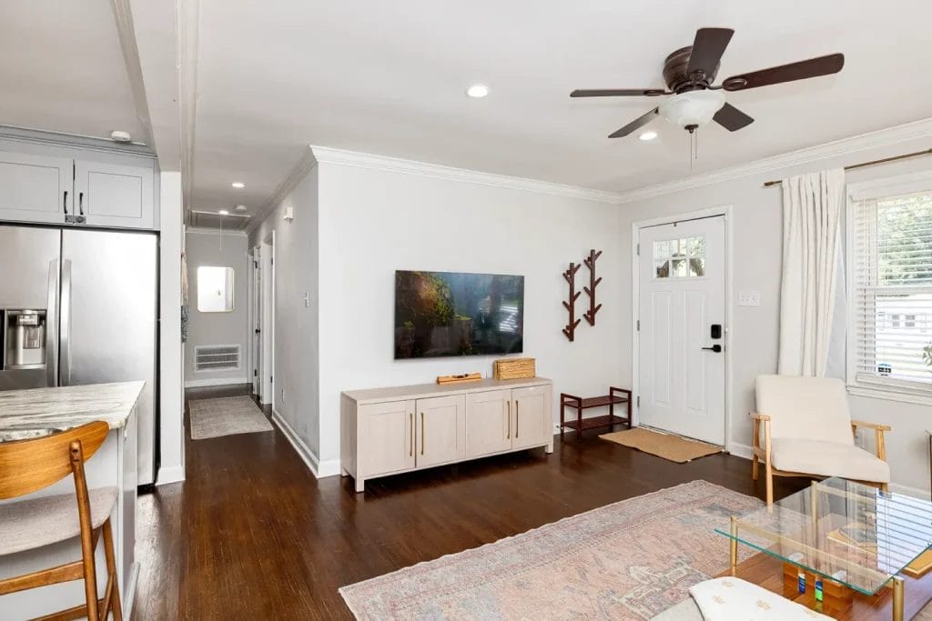A bright, modern living room with wood floors, a ceiling fan, a TV on a white console, and a glass coffee table. The space includes a kitchen area, a wooden chair, and a white front door with windows and curtains.