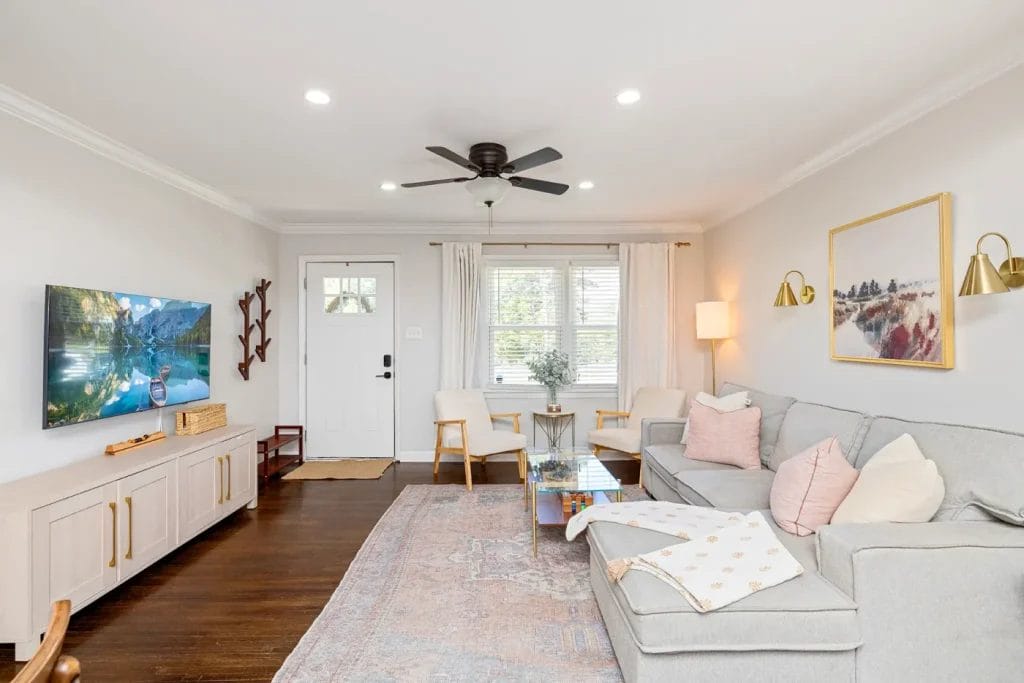 Bright living room with a gray sectional sofa, pastel pillows, a wall-mounted TV, a rug, two armchairs by the window, a ceiling fan, wall art, and neutral decor. White walls and dark wood floor complete the space.