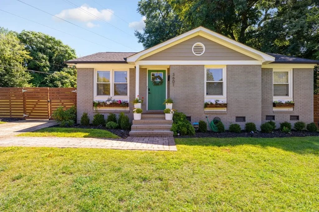 Single-story beige house with a green front door, flower boxes under the windows, potted plants by the steps, a tidy lawn, brick walkway, and a wooden fence in the background, on a sunny day.