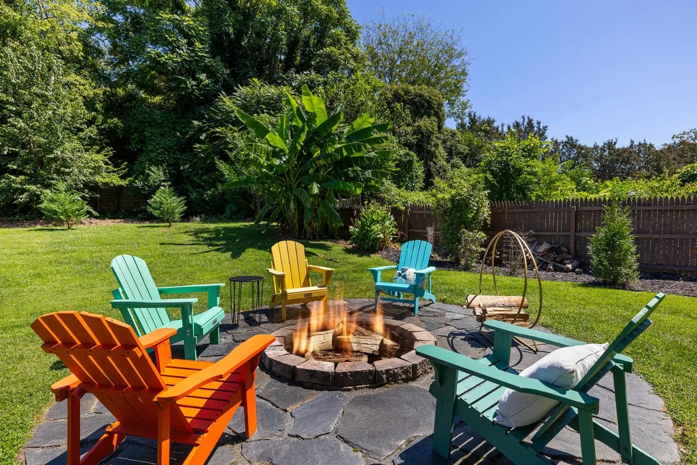 Six colorful Adirondack chairs are arranged around a stone fire pit in a grassy backyard, with lush green trees and plants in the background under a clear blue sky.