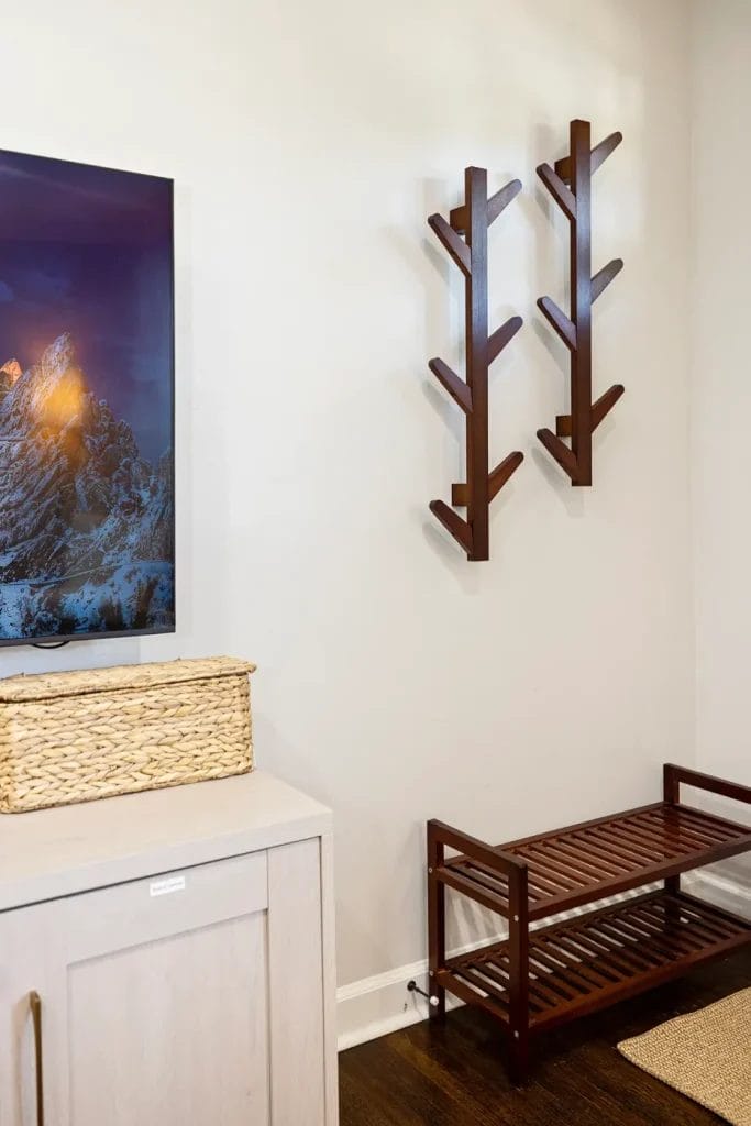 A modern entryway with a woven basket on a light cabinet, two wooden wall hooks shaped like branches, and a wooden shoe rack on a dark hardwood floor. A landscape photo is partially visible on the wall.