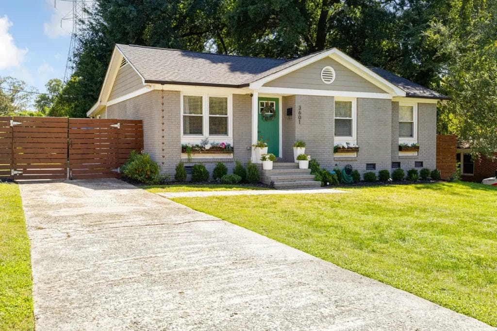 A small, single-story house with light brick exterior, green front door, and white-trimmed windows. Flower boxes are under the windows, and a concrete driveway leads to a wooden fence. The lawn is neatly trimmed.