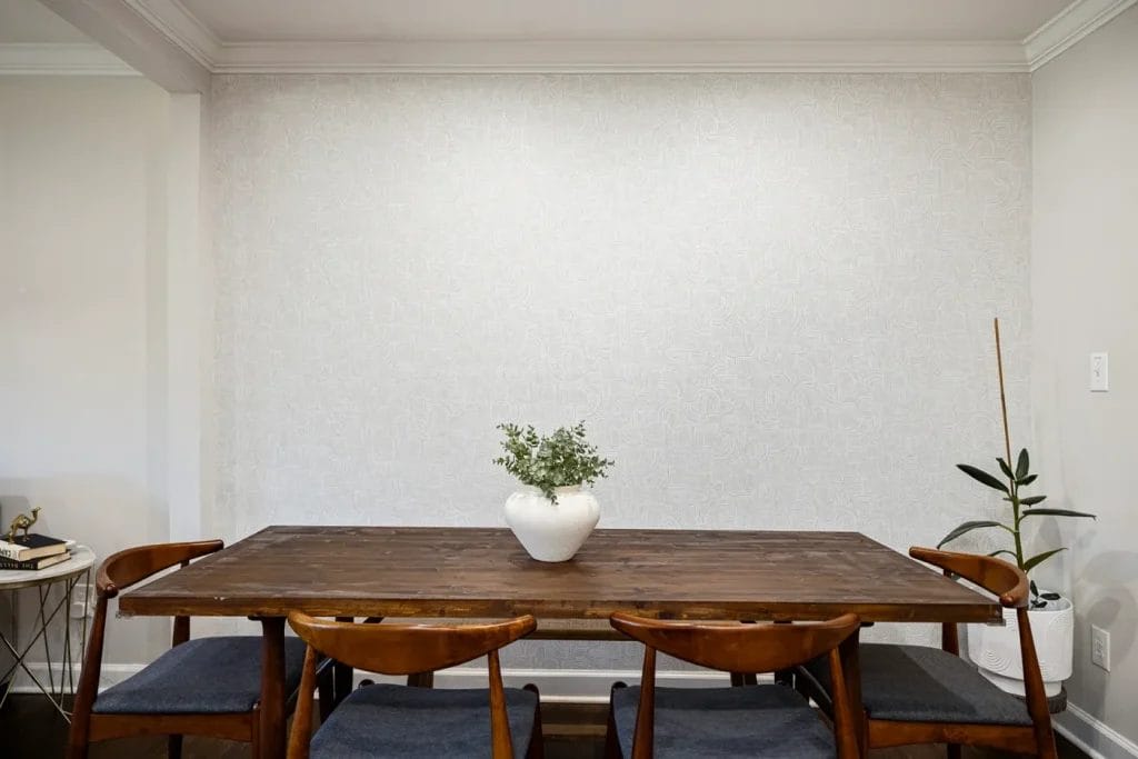 A wooden dining table with four chairs, a white vase with green foliage in the center, and a potted plant to the right, set against a plain light-colored wall.