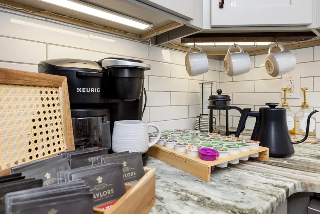 A kitchen coffee station with a Keurig machine, a French press, mugs hanging on hooks, various coffee pods organized in a tray, a black kettle, and tea sachets on a marble countertop.