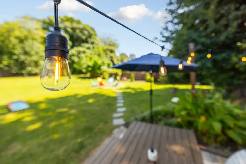 A close-up of a glowing outdoor string light hangs above a backyard with green grass, a path of stepping stones, a wooden deck, and a blue patio umbrella in the background.