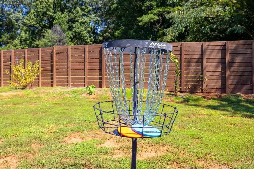 A disc golf basket with two discs inside stands on a grassy lawn, with a wooden fence and trees in the background under a sunny sky.