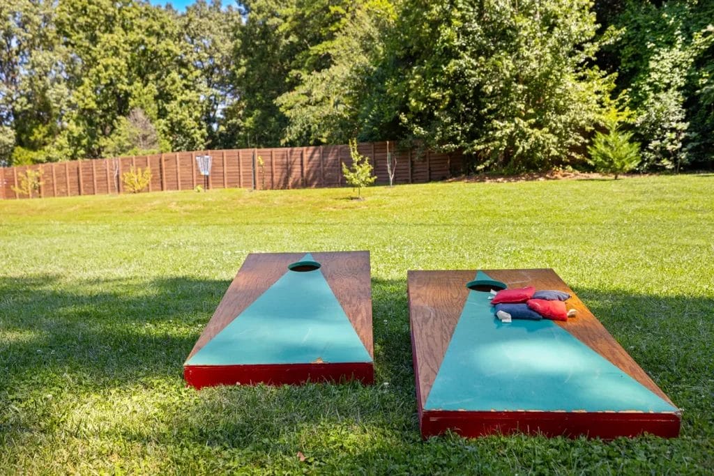 Two cornhole boards are set up on a grassy lawn, with bean bags placed on one board. Trees and a wooden fence are visible in the background on a sunny day.