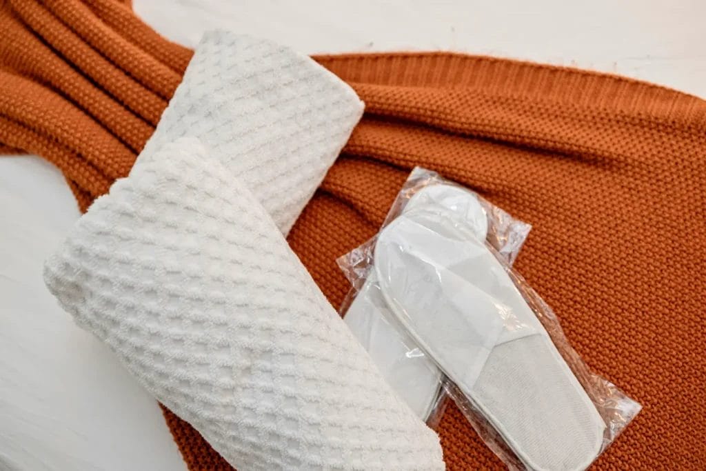 A pair of white textured slippers in plastic packaging rests on an orange knitted blanket, with a matching white waffle-texture bathrobe placed nearby on a light surface.