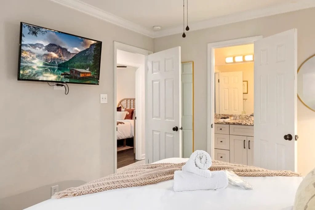 A cozy bedroom with a bed in the foreground, neatly folded towels on top, a wall-mounted TV showing a mountain scene, and open doors leading to a bathroom and another bedroom.