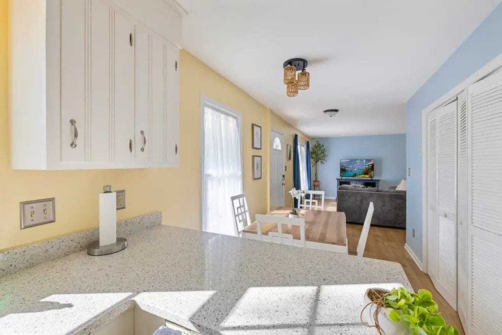 Bright kitchen with white cabinets and a granite countertop overlooks a dining area with a wooden table and white chairs. Sunlight streams through sheer curtains, and a cozy living room with a TV is visible in the background.