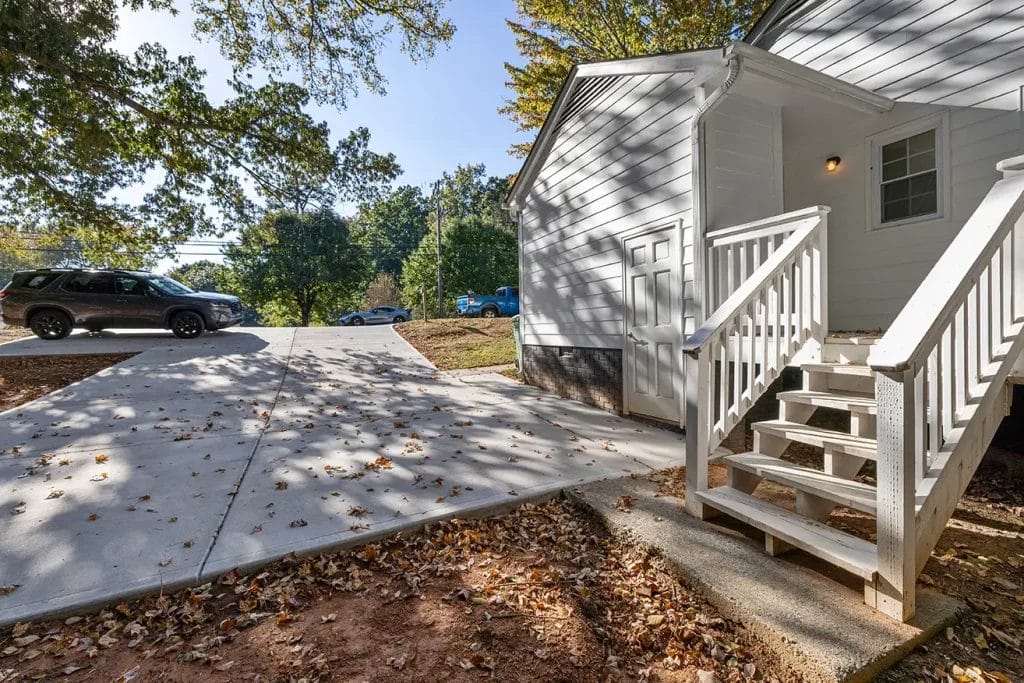 White wooden steps lead up to the side entrance of a house, next to a clean concrete driveway with scattered autumn leaves. Several cars are parked in the background under leafy trees.