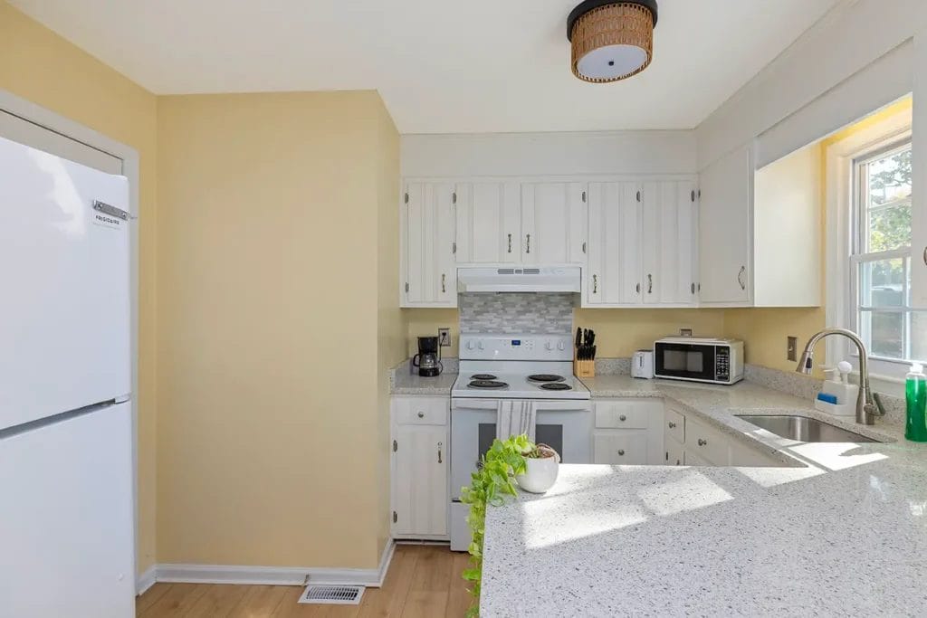 Bright kitchen with white cabinets, a white refrigerator, stove, and microwave. Sunlight streams through a window above the sink, illuminating the light countertops and a small potted plant on the island.