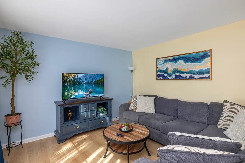 A cozy living room with a blue and gray sectional sofa, a round wooden coffee table, a TV displaying a lake scene, potted tree, and abstract wall art on pastel blue and yellow walls.