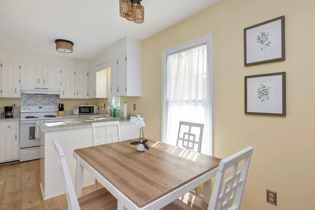 Bright kitchen and dining area with a wooden table, white chairs, white cabinets, appliances, and a window with sheer curtains. Two framed botanical prints hang on a light yellow wall.