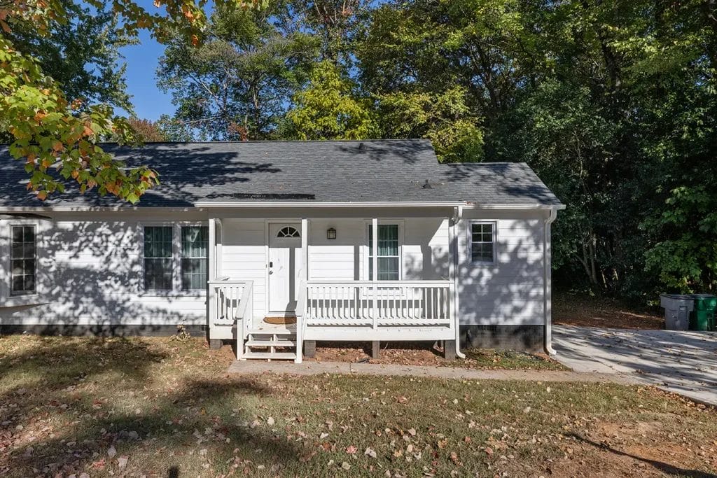 A small white house with a front porch and steps, surrounded by trees. The porch has a white railing, and the yard has patchy grass with some fallen leaves. The house is partially shaded by surrounding trees.