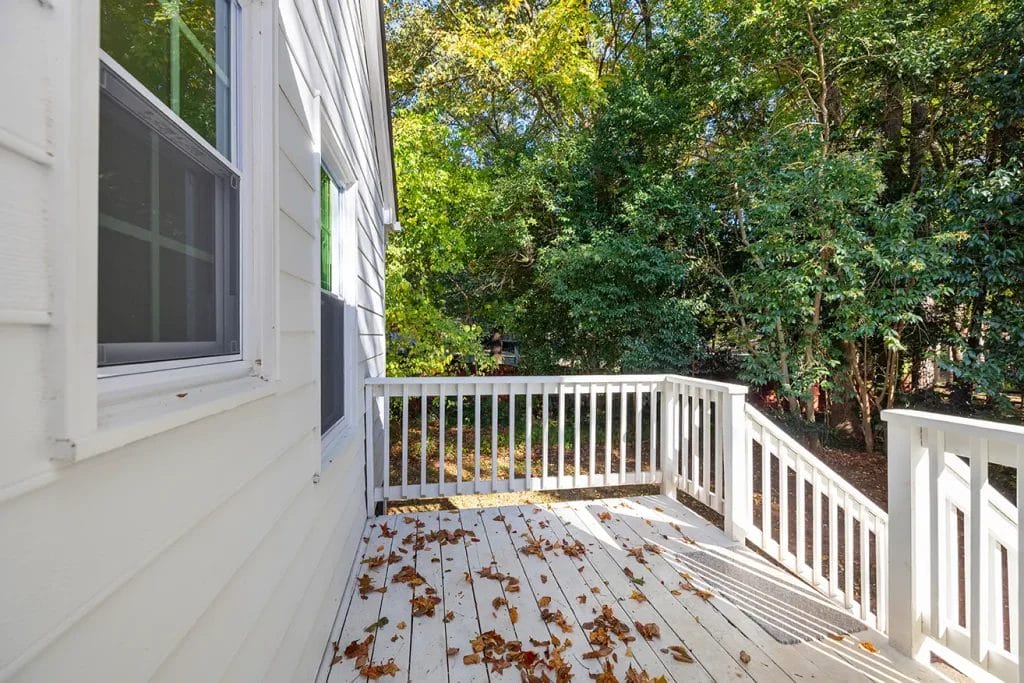 A white wooden deck with fallen leaves on the floor, attached to a white house, overlooks a backyard filled with green trees and shrubs on a sunny day.