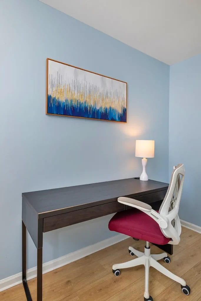A minimal home office with a black desk, a white desk chair with a red seat, a lit white lamp, light blue walls, and an abstract painting featuring blue, gold, and white tones above the desk.