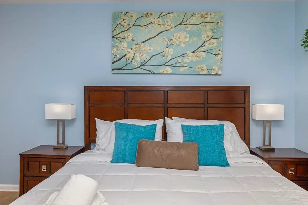 A neatly made bed with white linens, turquoise accent pillows, a brown bolster pillow, and dark wood headboard. Matching nightstands with modern lamps and a floral painting hanging on the light blue wall above.