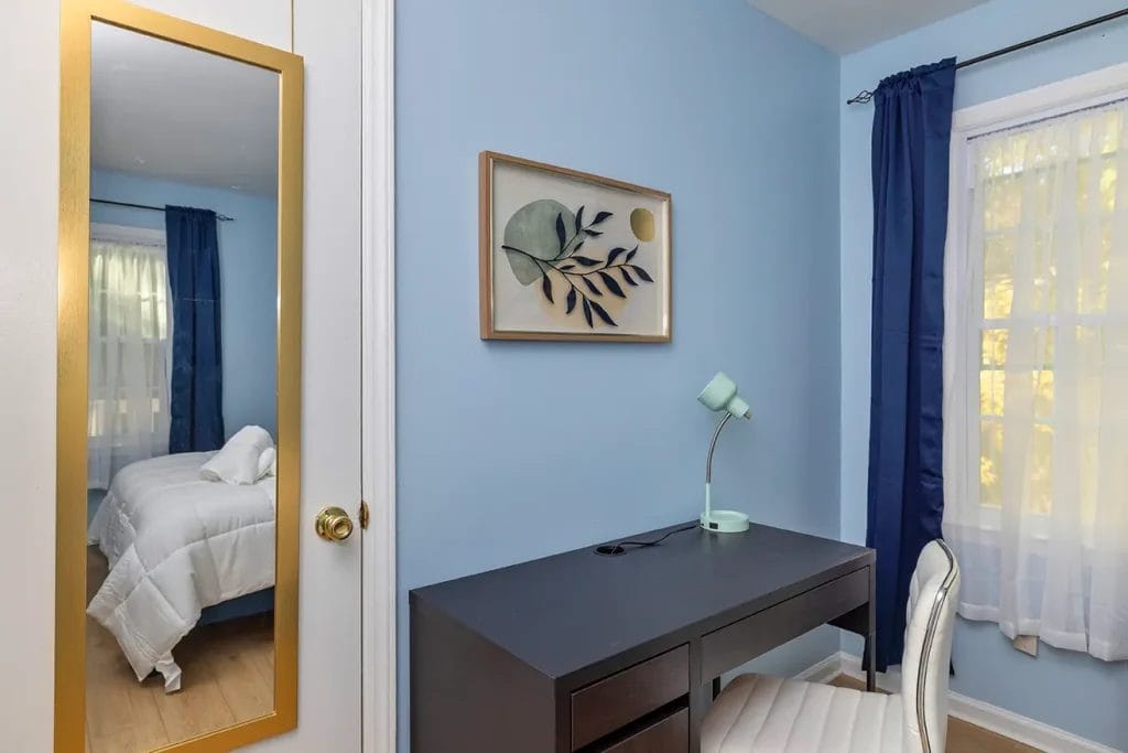 A bedroom corner with a blue wall, a black desk with a mint-green lamp, a white cushioned chair, a framed plant artwork, a full-length mirror, and windows with sheer and navy curtains. A bed is partially visible.