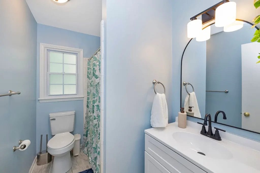 A clean, light blue bathroom with a white toilet, shower with a floral curtain, vanity with a black faucet, round mirror, white countertop, two towels on rings, and a window letting in natural light.