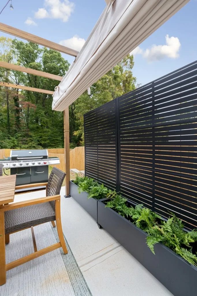 Modern outdoor patio with a wooden pergola, gas grill, dining table with woven chairs, privacy screen, and planter boxes filled with ferns; trees and a wooden fence in the background under a partly cloudy sky.