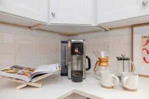 A modern kitchen counter with a coffee machine, two white mugs, a Chemex coffee maker, a French press, and an open cookbook on a stand under white cabinets.