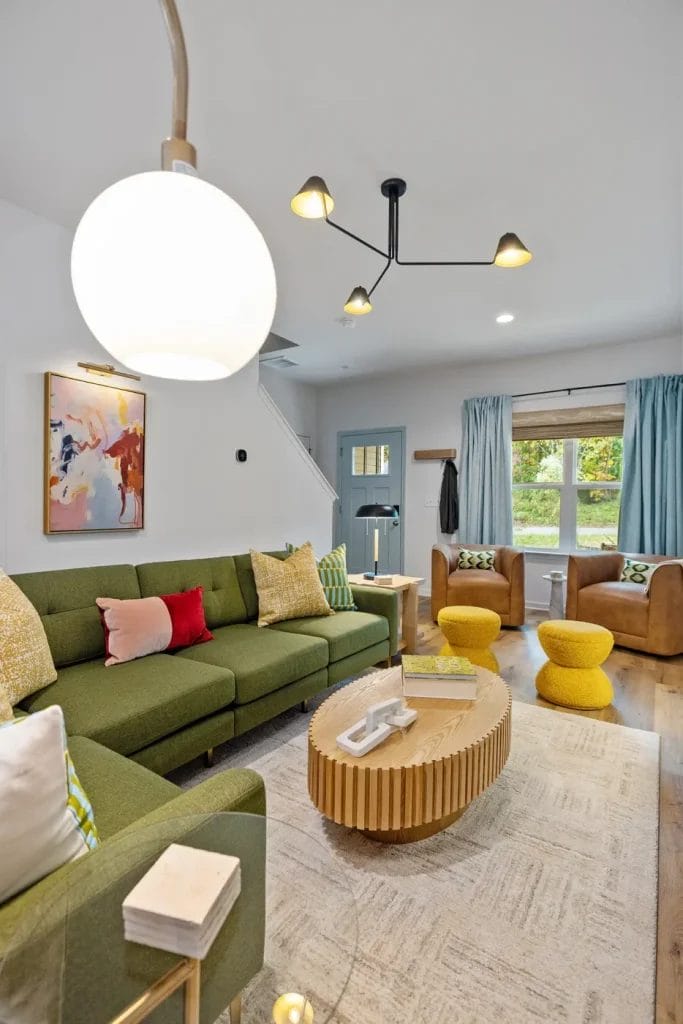 A modern living room with a green sectional sofa, colorful pillows, a round wooden coffee table, two yellow poufs, two brown armchairs, a large window with blue curtains, abstract wall art, and contemporary lighting.