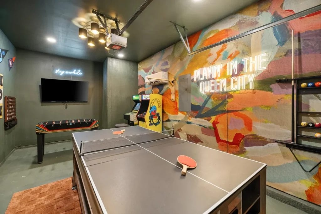 A colorful game room featuring a ping pong table, foosball table, arcade machine, wall-mounted TV, neon “stay wild” sign, and a vibrant mural with the words "PLAYIN' IN THE QUEEN CITY" in glowing letters.