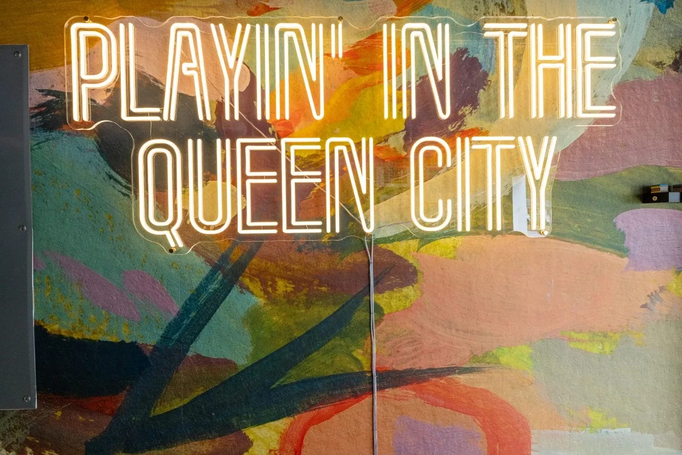 Neon sign reading "PLAYIN' IN THE QUEEN CITY" is mounted on a colorful, abstract painted wall with bold brushstrokes and vibrant hues in the background.