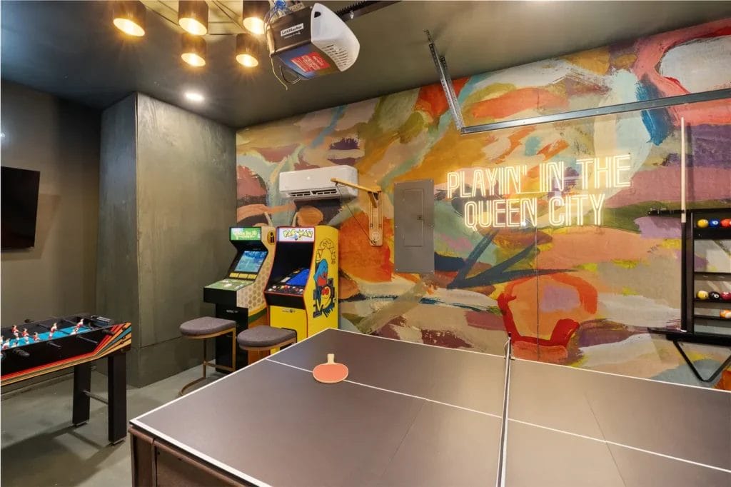A colorful game room featuring a ping pong table, foosball, two arcade machines, and a neon sign reading “PLAYIN’ IN THE QUEEN CITY” on a vibrant, abstract mural wall.