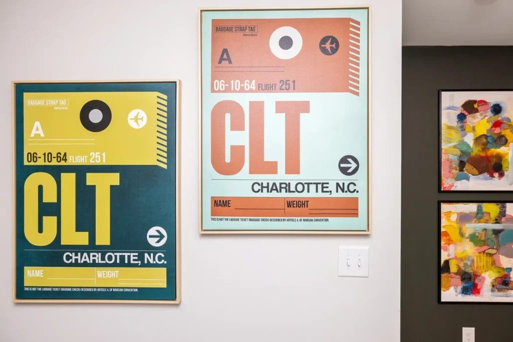 Two framed posters inspired by vintage baggage tags read "CLT, Charlotte, N.C." in bold letters. Next to them are two abstract, colorful art pieces on the wall. Light switches are below the posters.
