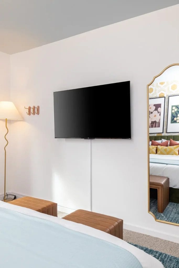 A modern bedroom with a wall-mounted TV, a large gold-framed mirror, two brown stools, a floor lamp with a wavy stand, and framed art reflected in the mirror.