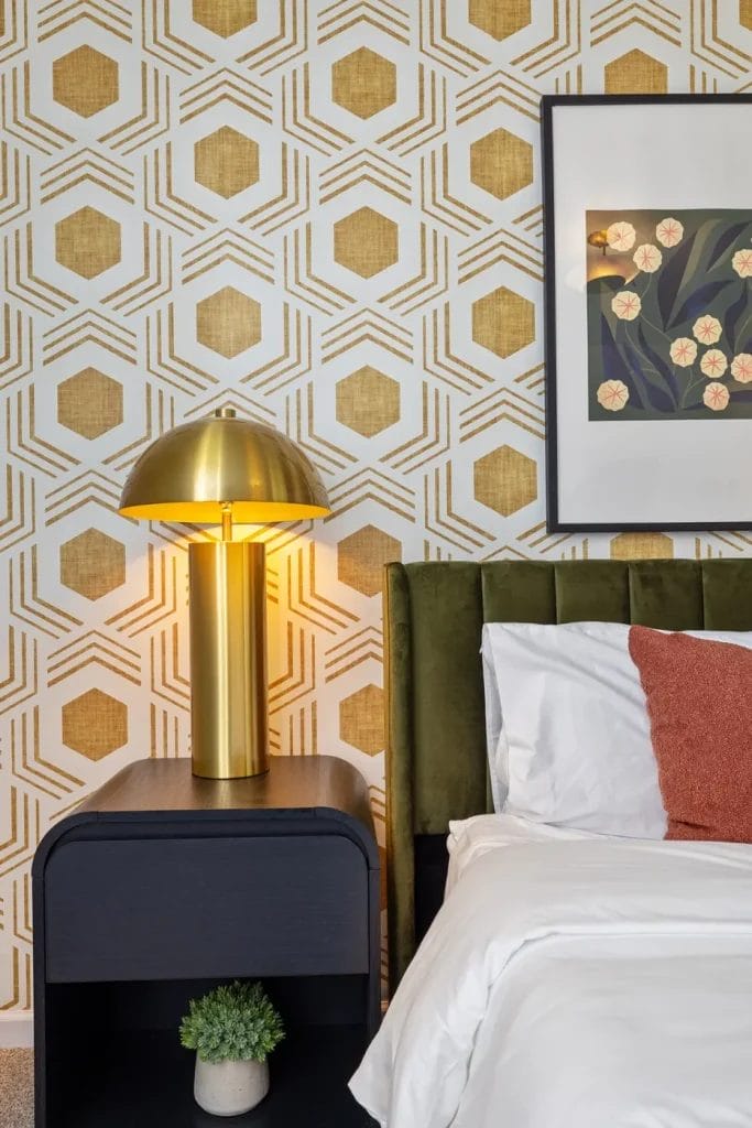 A modern bedroom features a gold geometric wallpaper, a gold dome lamp on a black nightstand, a green velvet headboard, white bedding with a rust pillow, a framed floral print, and a small potted plant.