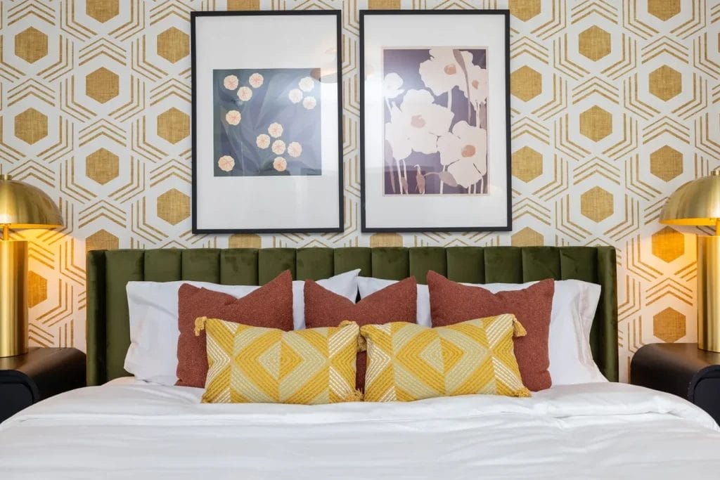 A stylish bed with mustard yellow and rust pillows and a green velvet headboard, flanked by gold lamps. Behind, geometric wallpaper and two floral art prints decorate the wall.