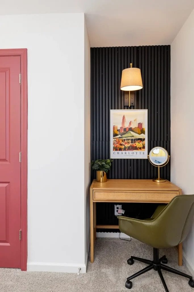 A small home office nook with a wooden desk, green swivel chair, round mirror, potted plant, and a wall lamp above a framed Charlotte print, set against a black slatted accent wall and a red door on the left.