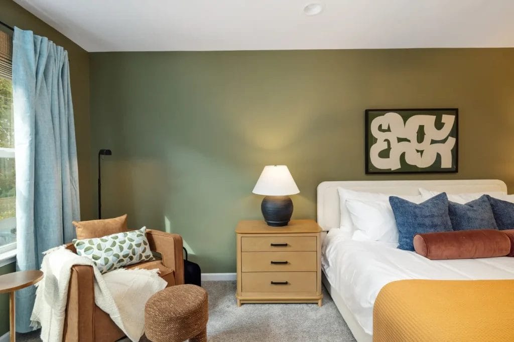 A cozy bedroom with an olive green accent wall, a modern bed with white linens and blue pillows, a wooden nightstand with a lamp, an abstract artwork, and a brown armchair by a window with blue curtains.
